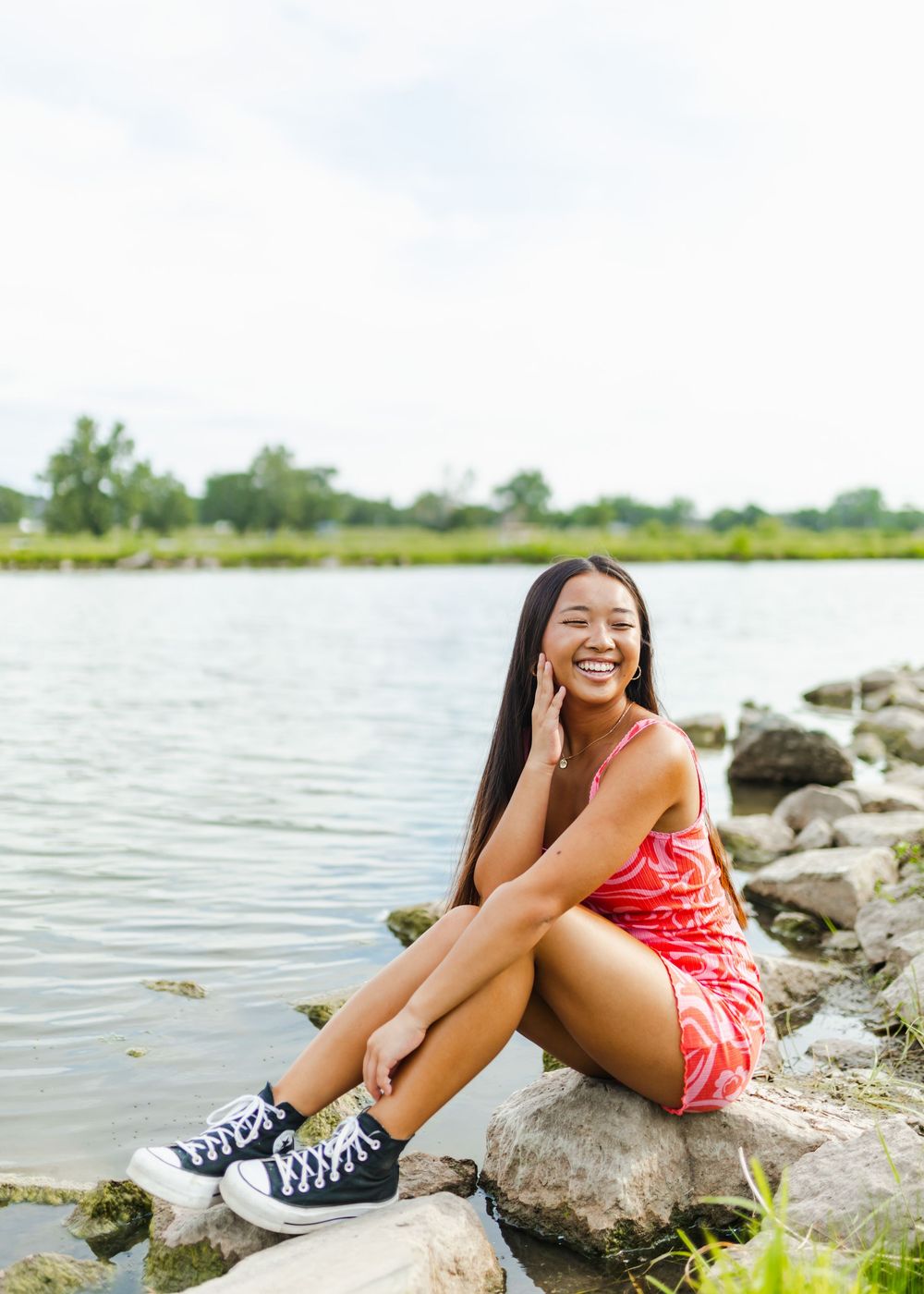 Person in a red dress and sneakers sits on rocky lakeside shore during a sunny summer day.