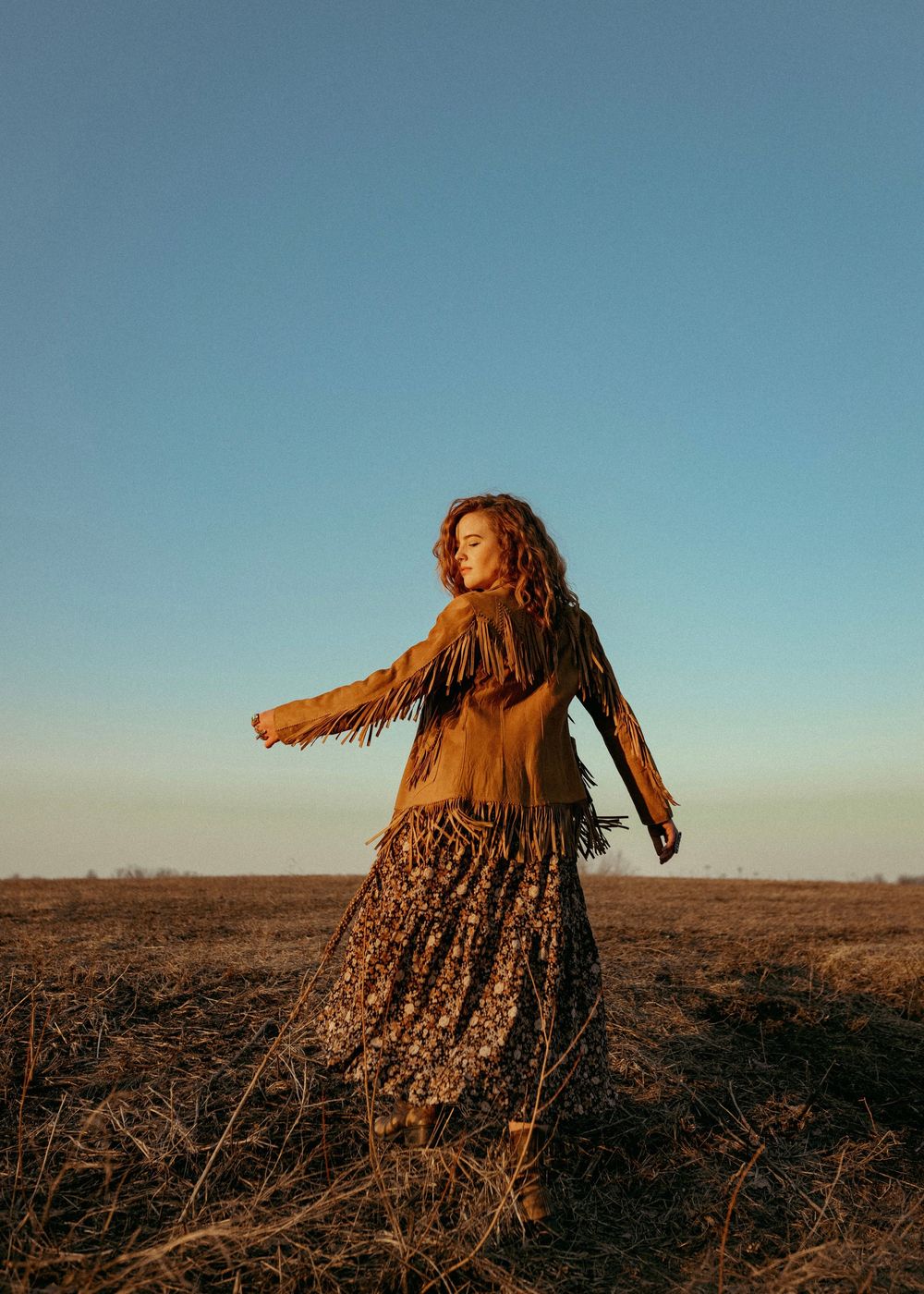 A person in a brown dress and jacket twirls in a golden field at sunset against a clear blue sky.