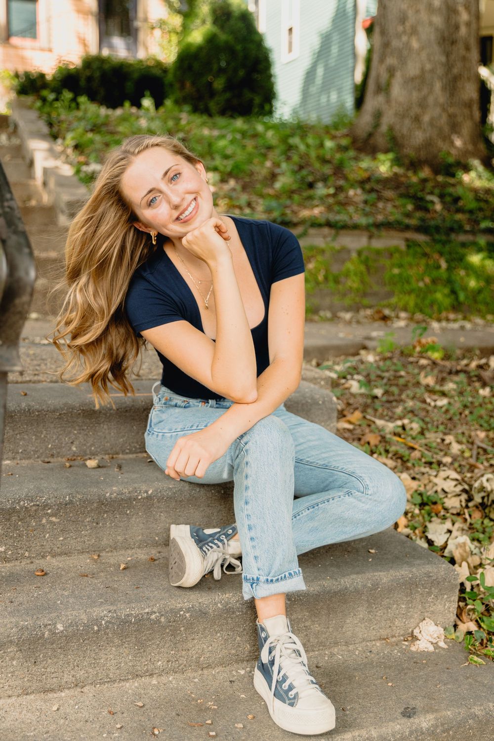 A person in a navy t-shirt and jeans sits casually on concrete steps outside while smiling on a sunny day.