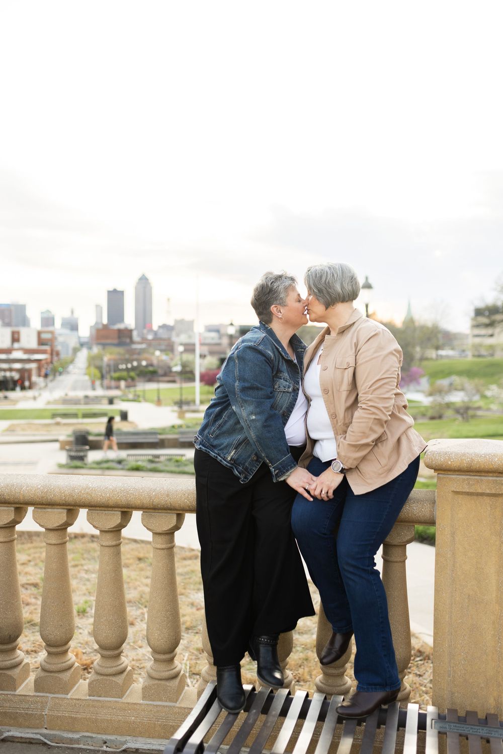 A romantic couple shares a kiss on a stone balcony overlooking the Des Moines city skyline on an overcast day.