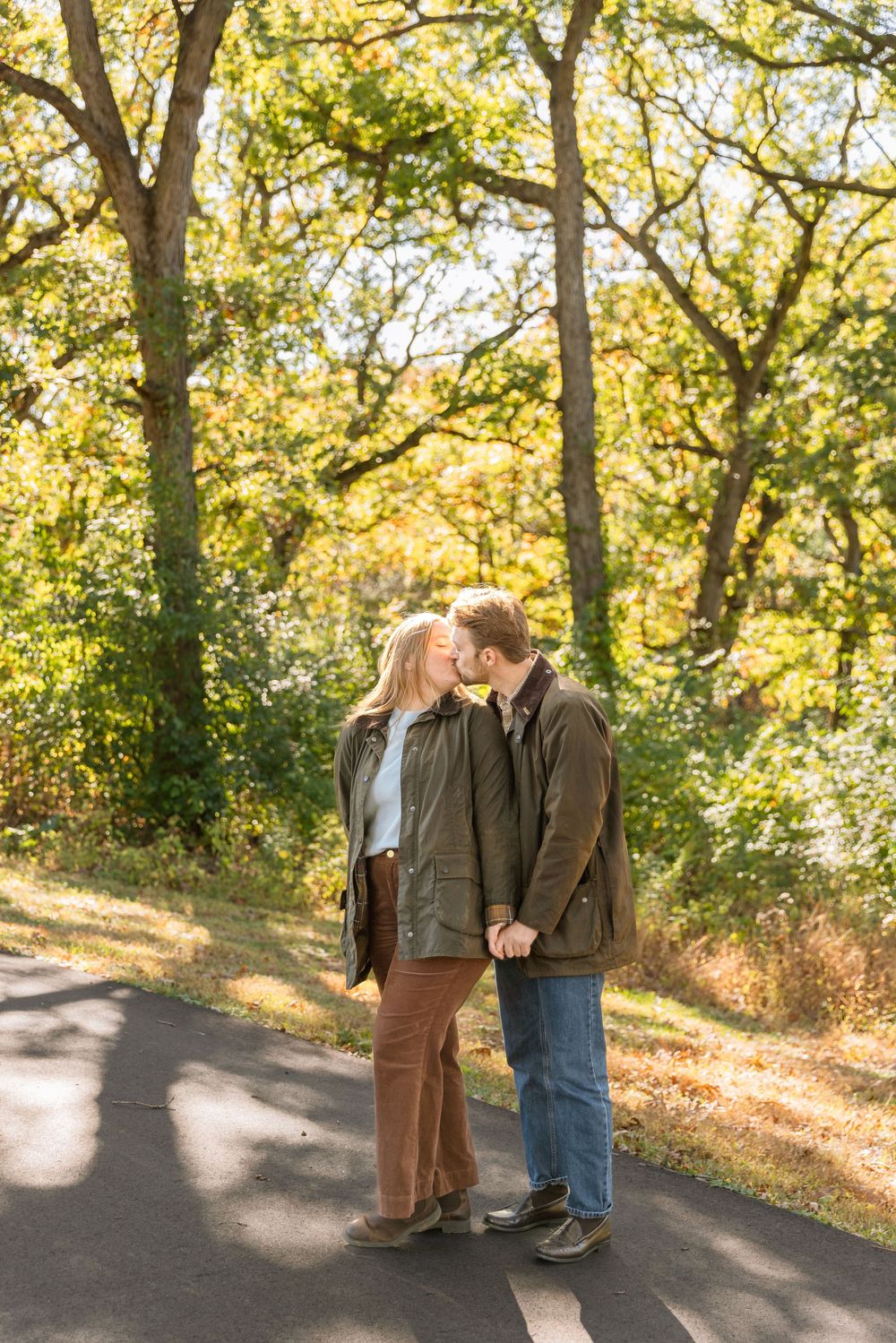 A couple shares a tender moment on a sunlit autumn path surrounded by golden trees and falling leaves.