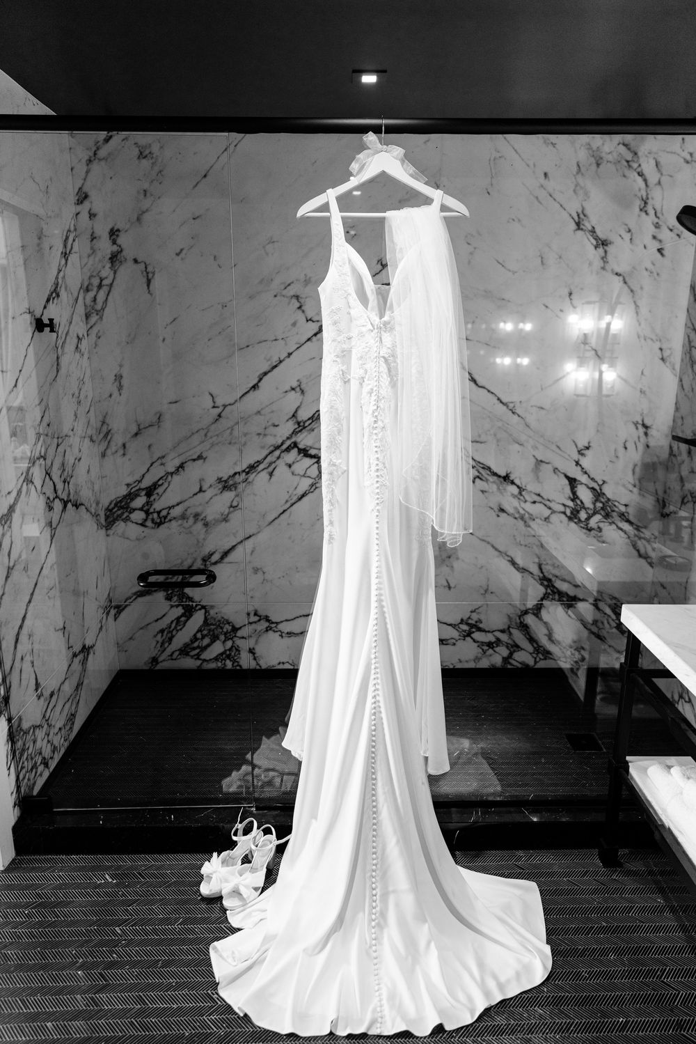 An elegant white wedding gown with lace details hangs on display against a marble wall with shoes beneath it.
