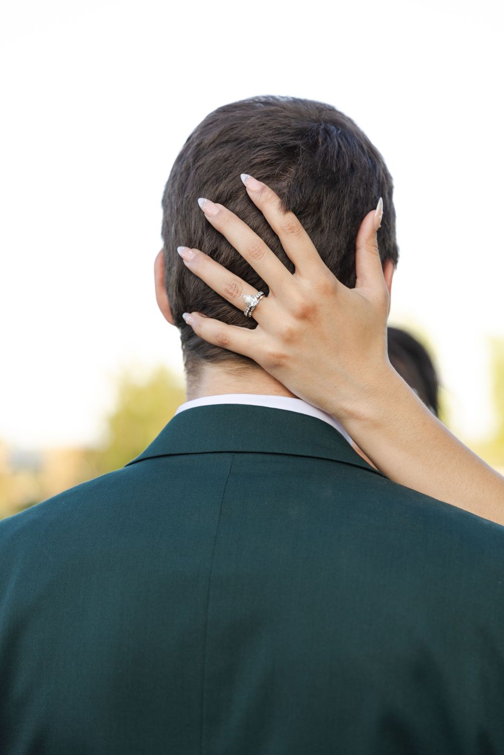 A hand with painted nails and rings rests against the back of someone's head wearing a dark green suit jacket.