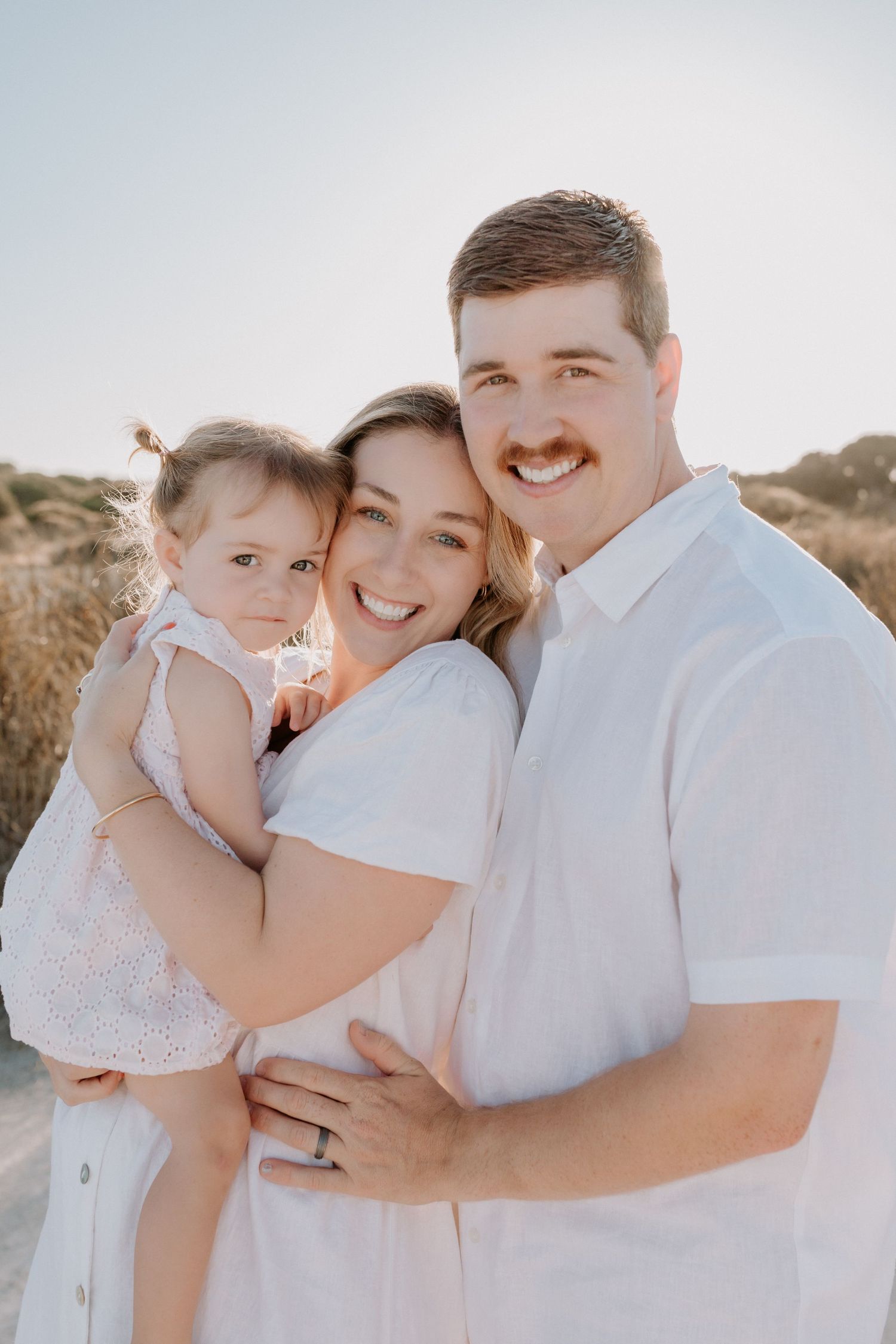 A loving family poses together outdoors in white outfits during a sunny photoshoot against a natural desert background.