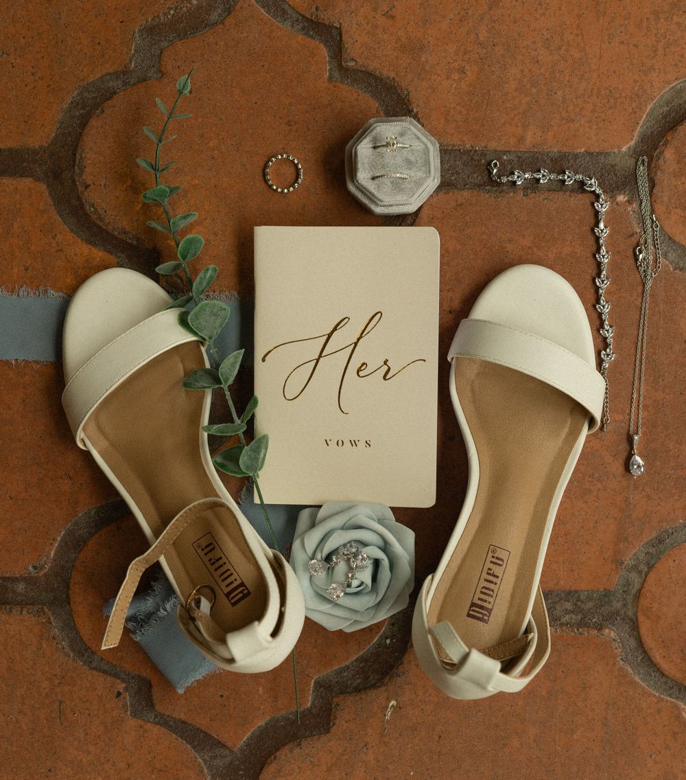 Elegant white bridal sandals arranged with a 'Her' vows book and eucalyptus on terracotta tiles.