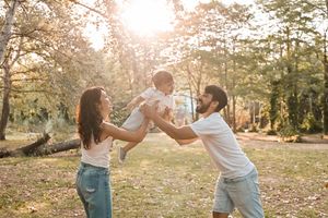 Happy young family playing with baby in sunny park, parents lifting toddler outdoors in golden light.