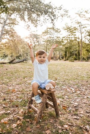 Joyful toddler sitting on rustic wooden stool outdoors, arms raised in excitement, wearing white shirt and blue shorts.