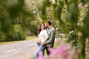 Couple sharing a tender moment on a garden path surrounded by lush greenery and pink roses during maternity photo shoot.