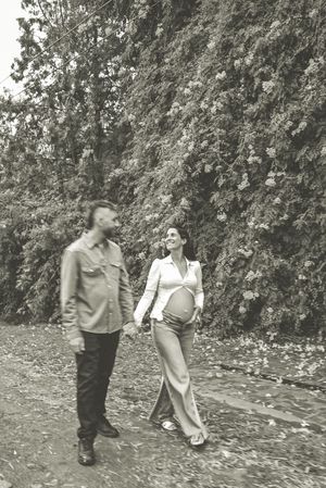 Black and white photo of a pregnant couple holding hands and walking outdoors near a lush ivy-covered wall.