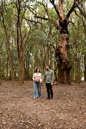 Pregnant couple holding hands in a lush forest, standing near a large ancient tree surrounded by fallen leaves.