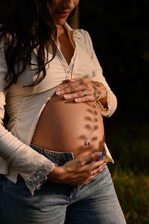 Pregnant woman in white shirt and jeans cradling bare baby bump outdoors with leaf shadow pattern in golden sunlight.