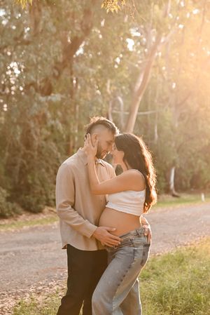 Couple sharing a tender moment outdoors, woman with baby bump in white crop top and jeans, golden sunset light filtering through trees.