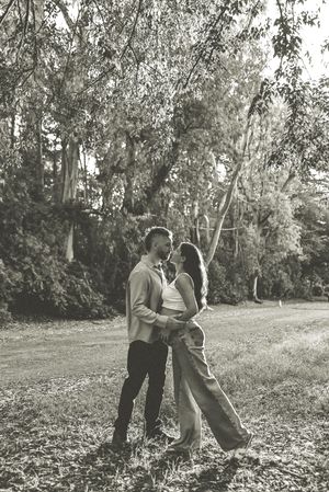 Black and white photo of a romantic couple embracing and almost kissing outdoors among lush trees in a park.