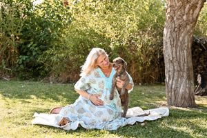 Pregnant woman in floral dress sits on blanket outdoors, lovingly interacting with a brown puppy near a tree.