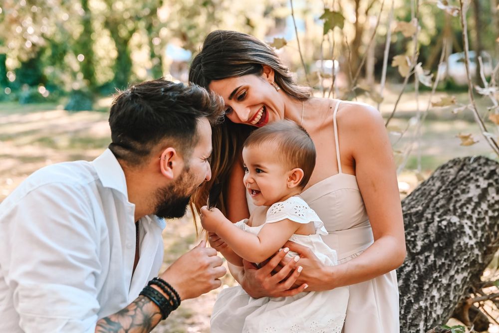 Happy young family with tattooed father, smiling mother in white dress, and laughing baby enjoying outdoors in sunny park.