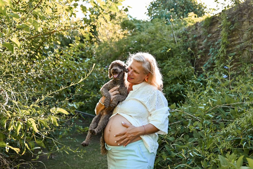 Pregnant woman with blonde hair holds a small dog outdoors among lush green foliage.