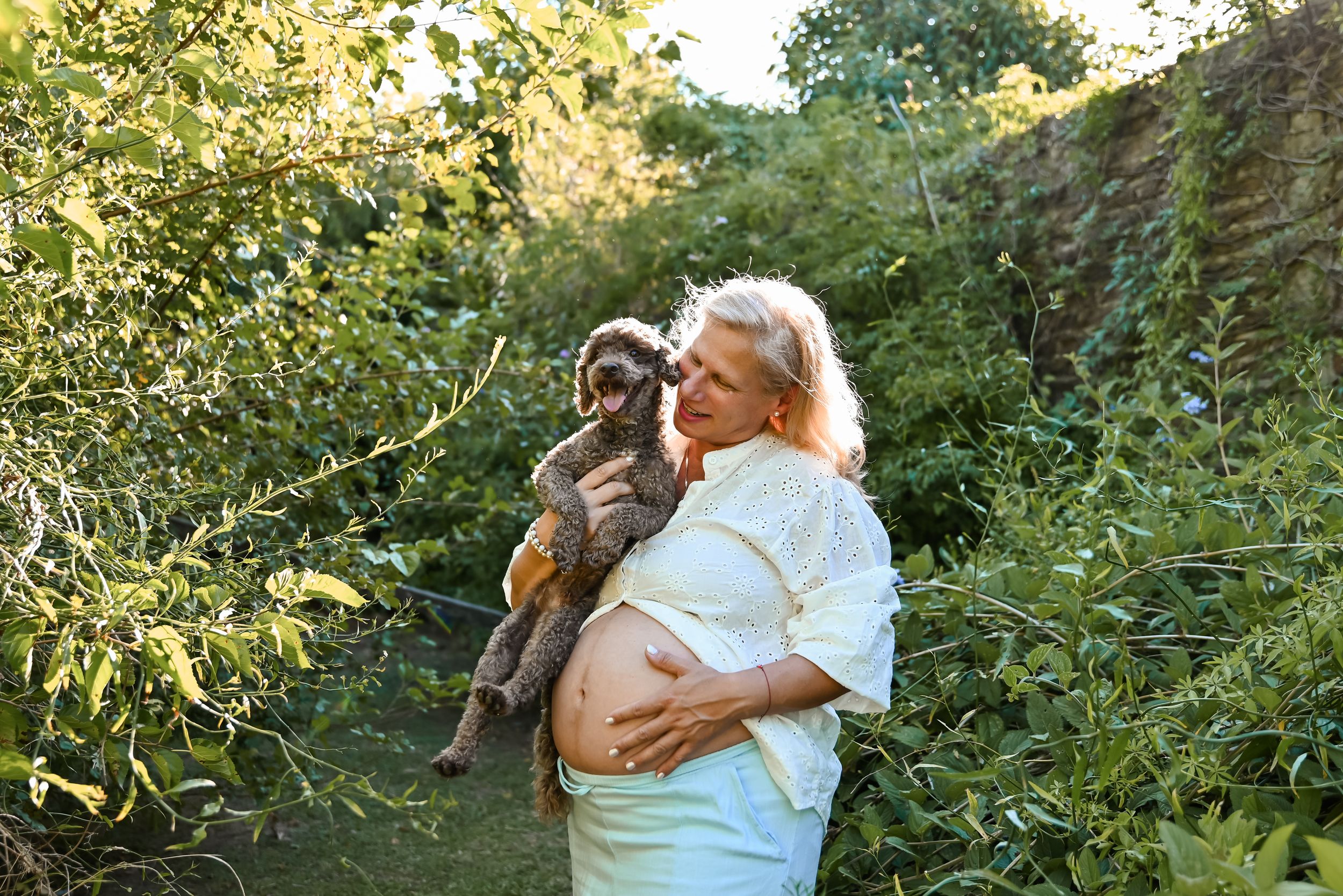 Pregnant woman with blonde hair holds a small dog outdoors among lush green foliage.