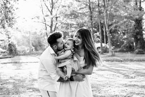 Black and white photo of happy family with parents kissing and hugging their laughing toddler outdoors.