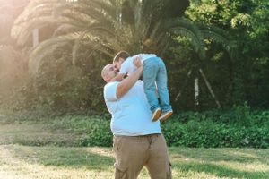 Parent lifting child playfully in garden with palm trees at sunset.