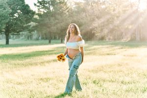 Expectant mother holding sunflowers in golden sunlit field.
