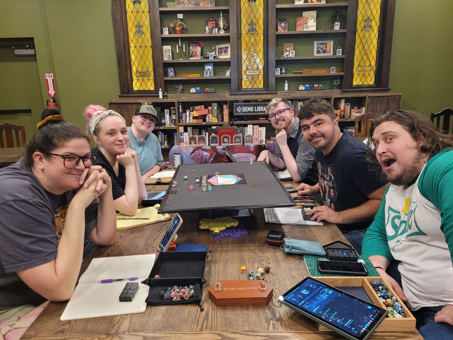 six individuals smiling surrounding ttrpg gaming table with stained glass shelves in the background