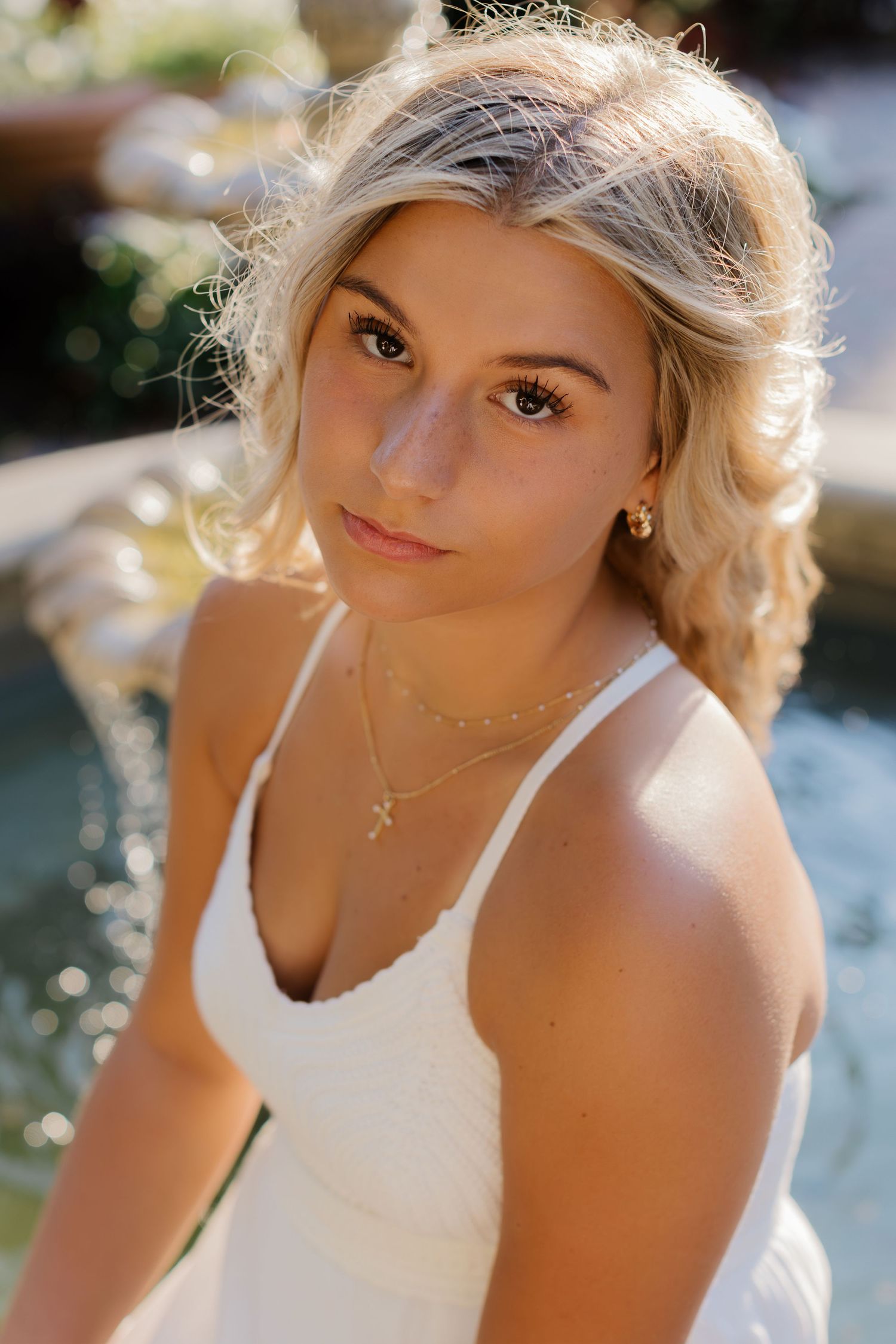 Portrait of blonde woman wearing white top and delicate necklace in outdoor setting.