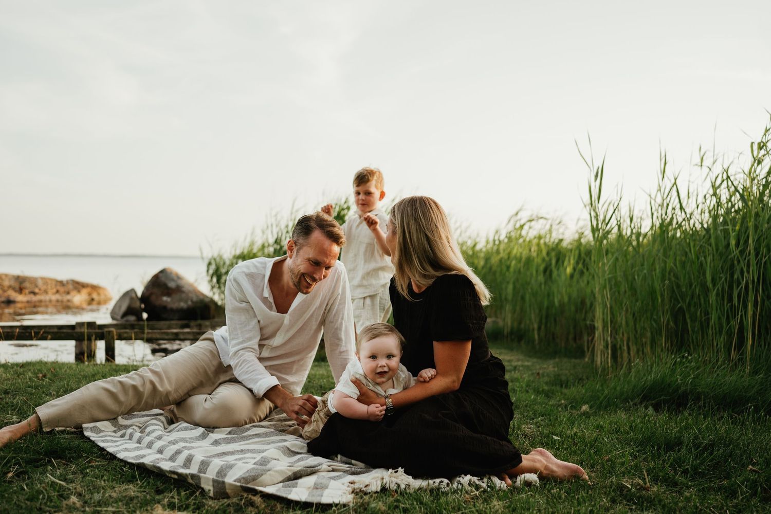 Family relaxing together on a picnic blanket by the water with tall reeds in background.