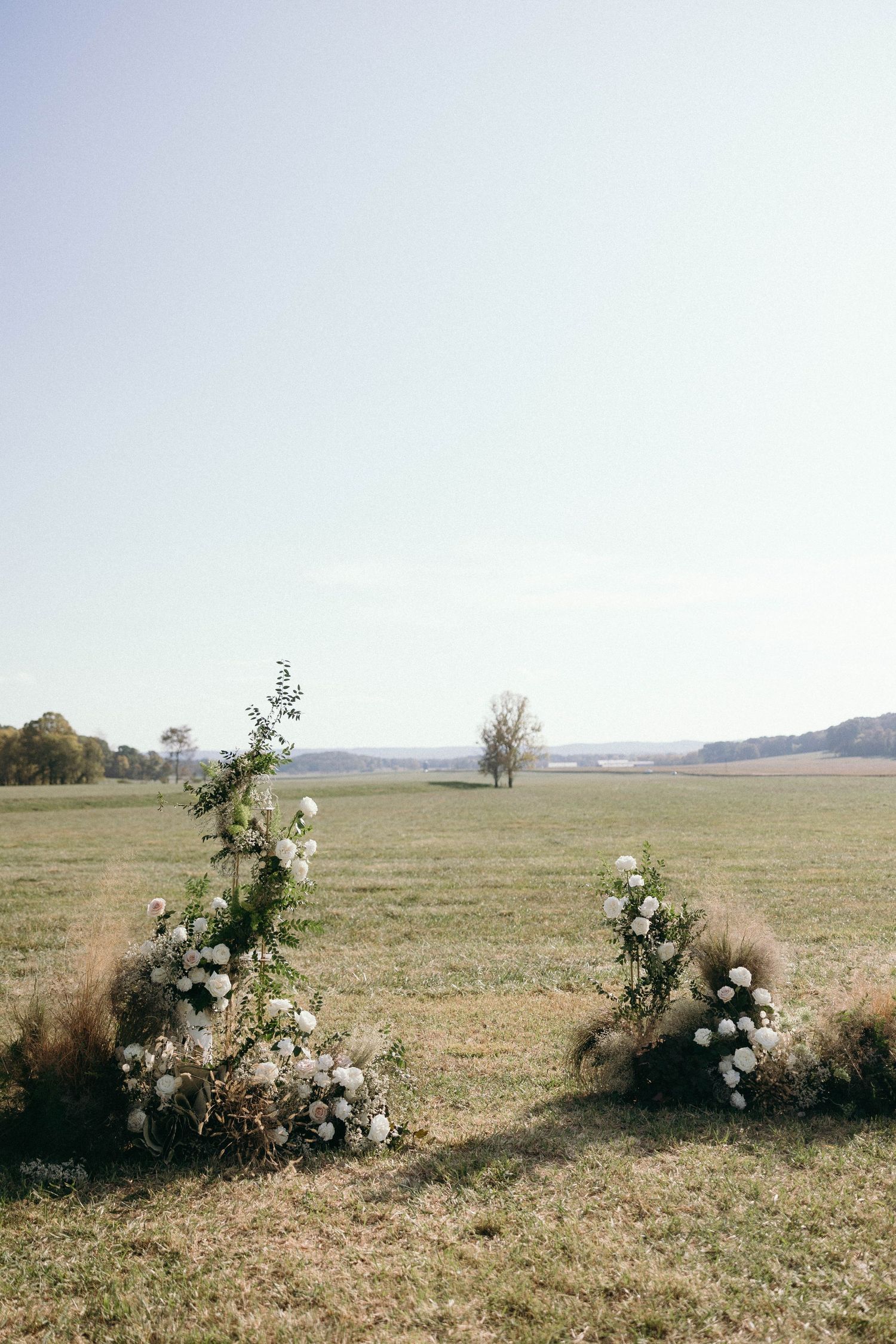 White floral arrangements create an elegant outdoor display in a grassy field with rolling hills in the background.