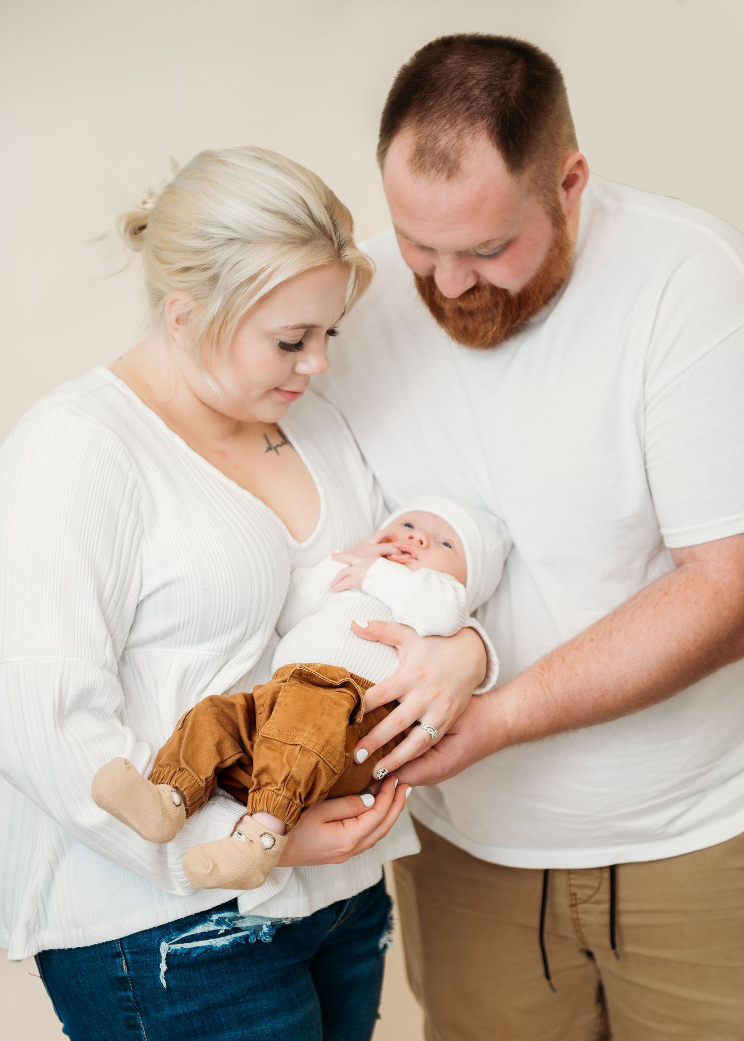 Couple in matching white shirts gently holding their newborn baby wrapped in brown blanket.