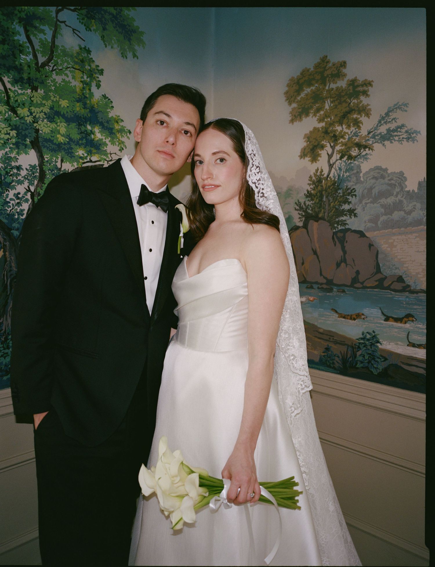 Classic wedding portrait with couple in formal attire posing against scenic wallpaper backdrop with lily bouquet.