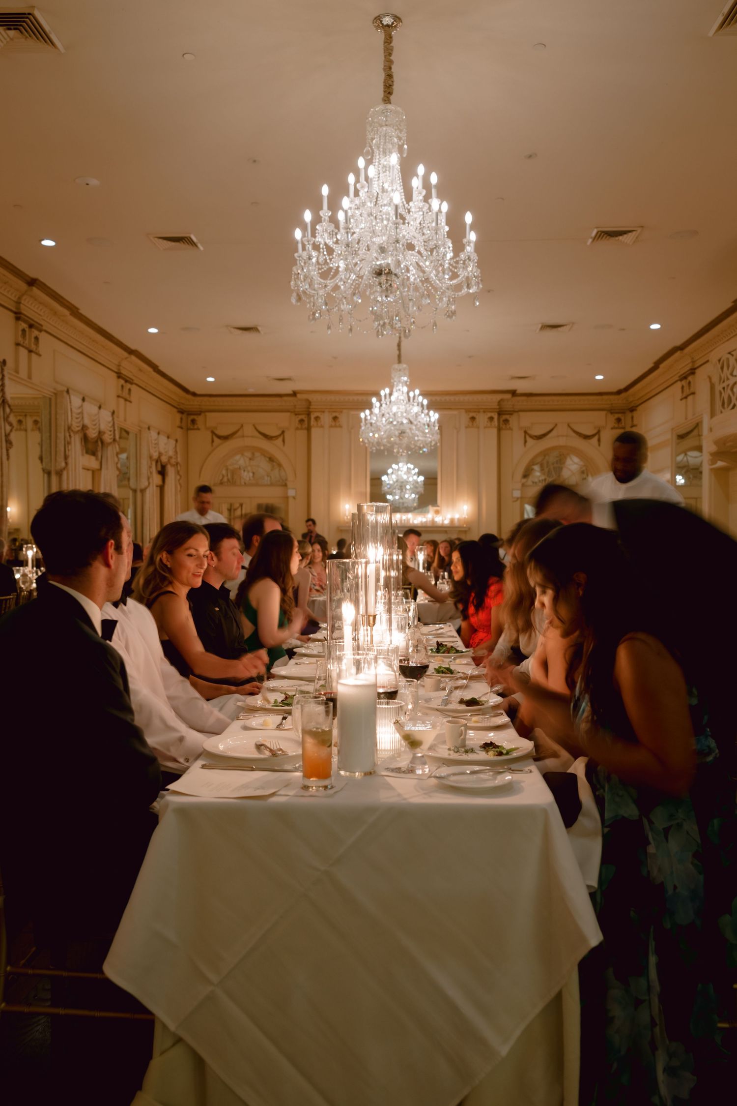 Elegant dining room with crystal chandeliers and long table set for formal dinner with candlelight and white linens.