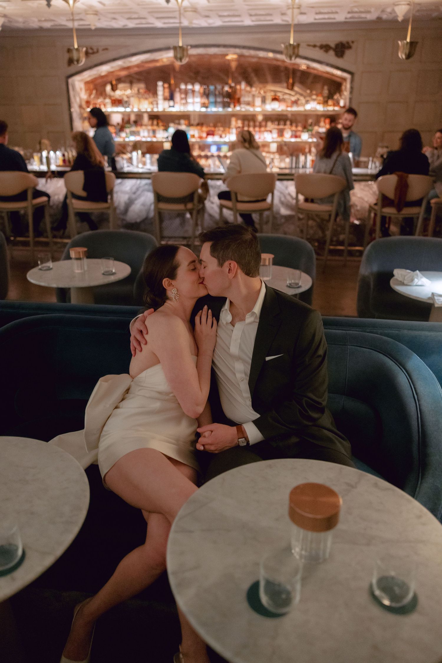 Romantic couple sharing an intimate kiss at an upscale bar with marble tables and blue velvet seating.