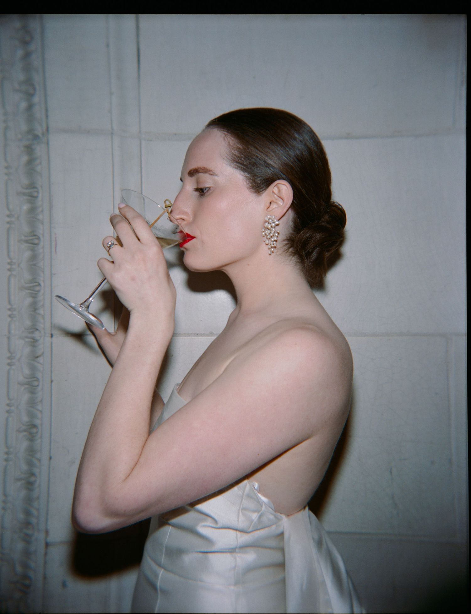Profile view of someone in an elegant white gown sipping from a champagne glass with sparkly earrings in vintage film style.