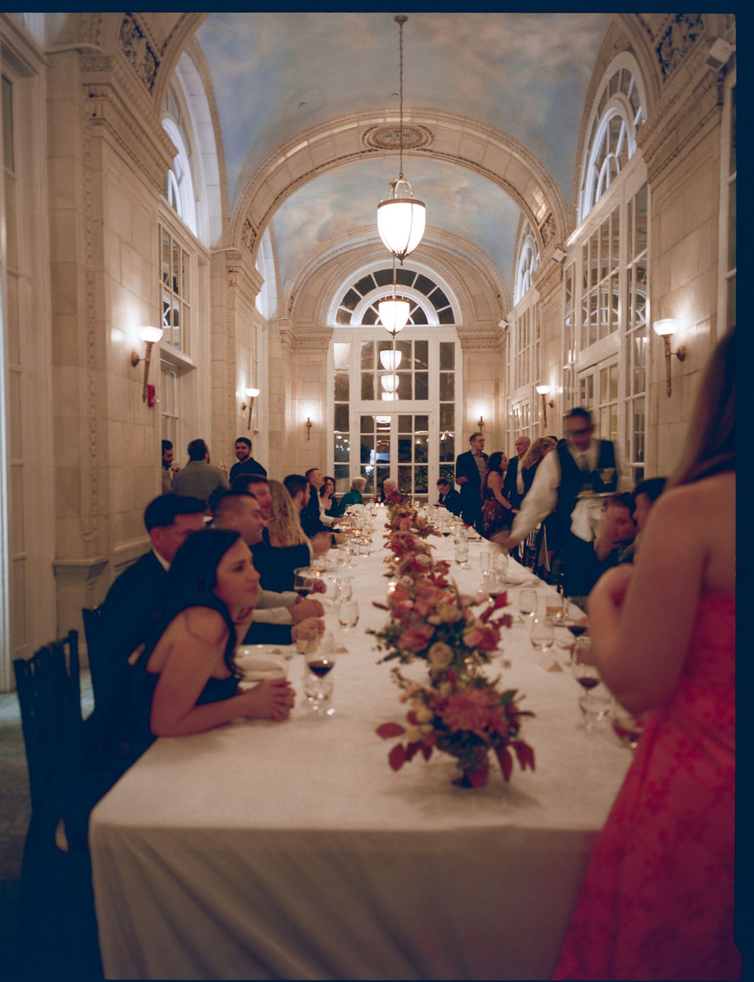 Elegant wedding reception with long table setting under arched hallway with floral centerpieces and ornate lighting.