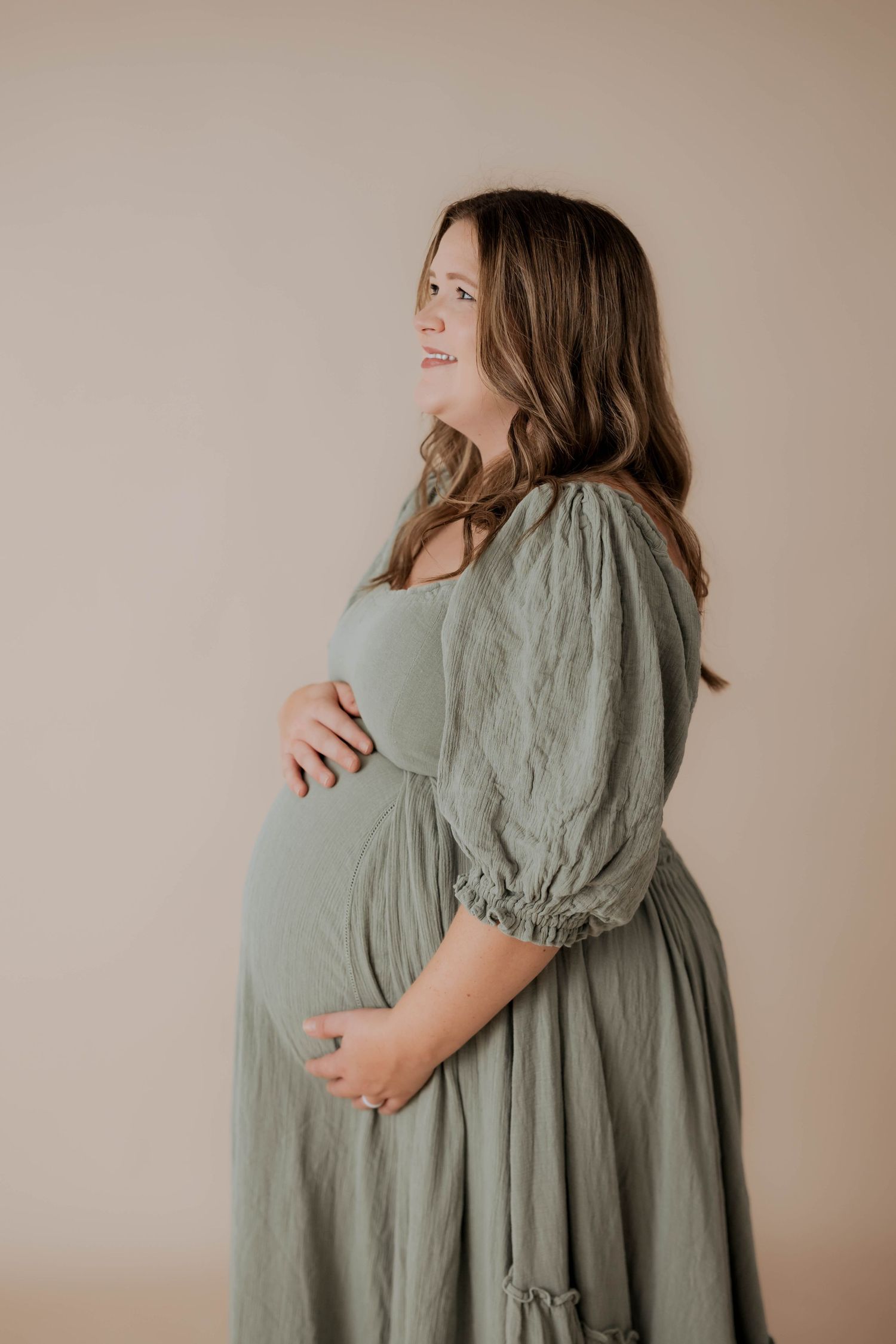 Expectant mother in sage green dress poses for maternity portrait against beige backdrop.