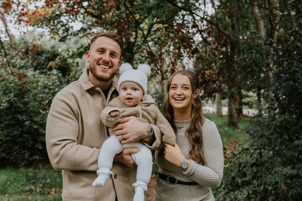 A family of three poses outdoors in matching beige and white outfits surrounded by lush greenery on a fall day.