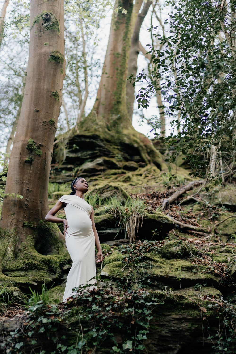 A woman in a white gown poses gracefully among moss-covered rocks and tall trees in a lush forest setting.