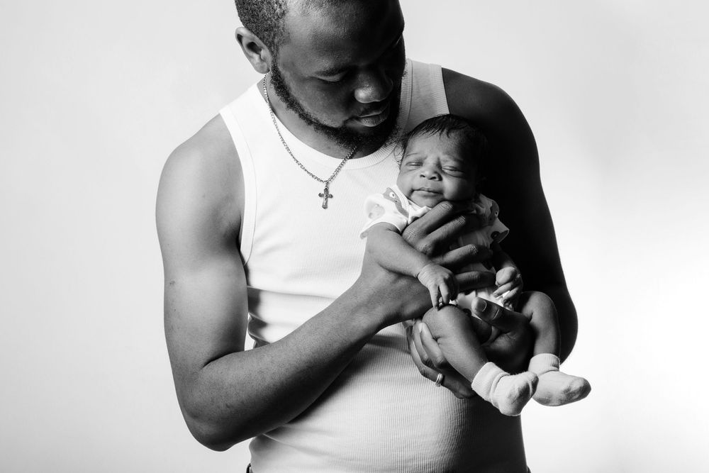 Black and white photo of a muscular man in a white tank top tenderly holding a newborn baby close to his chest.