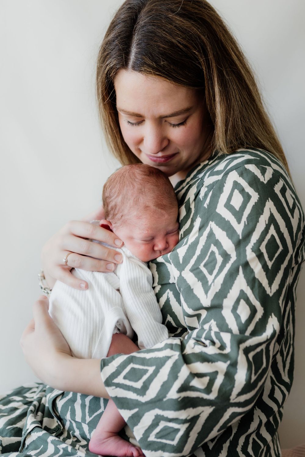 Mother in geometric print sweater cuddles sleeping newborn baby against white background.