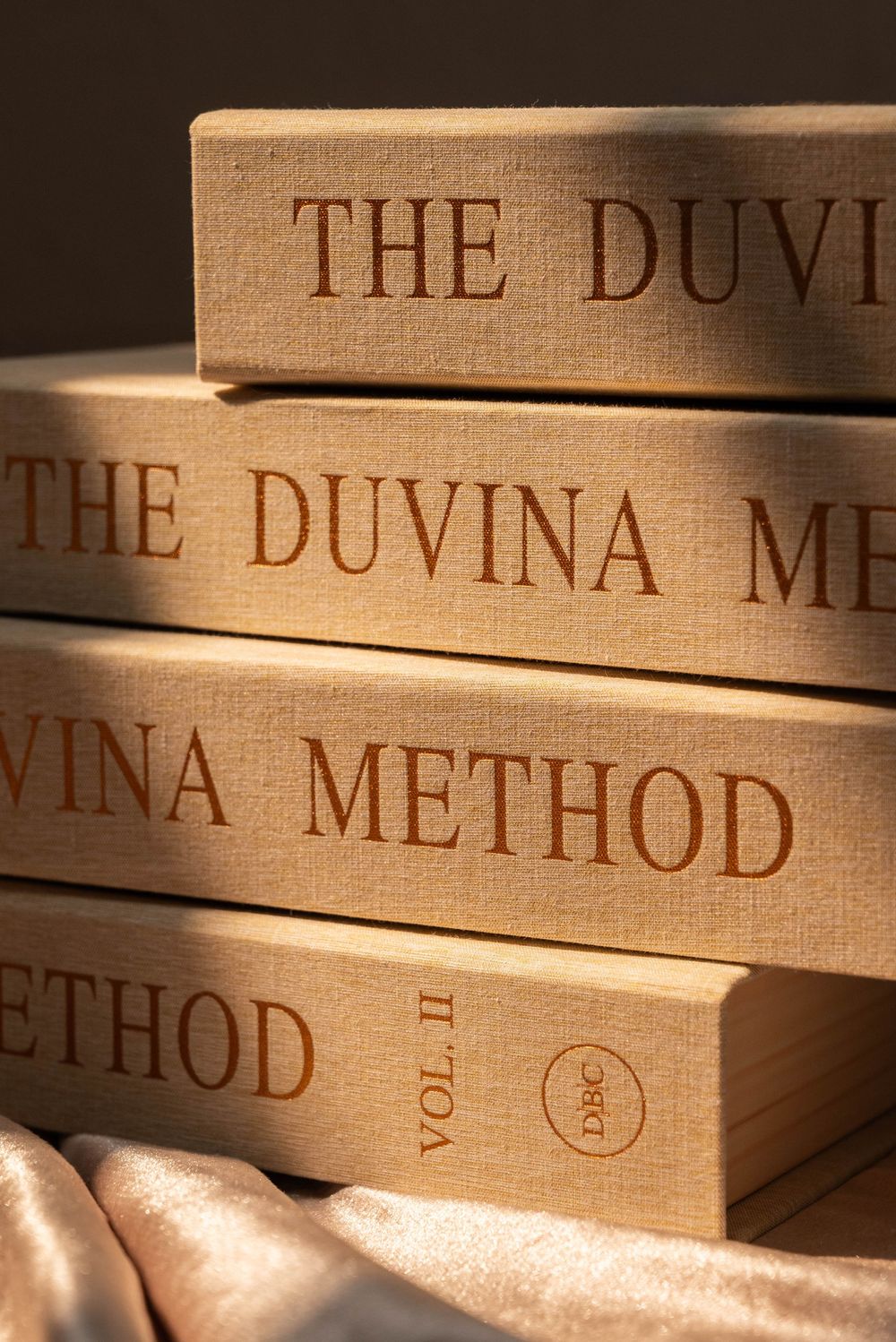 Stack of four beige linen-bound books titled The Duvina Method, Vol. II, arranged on silk fabric.