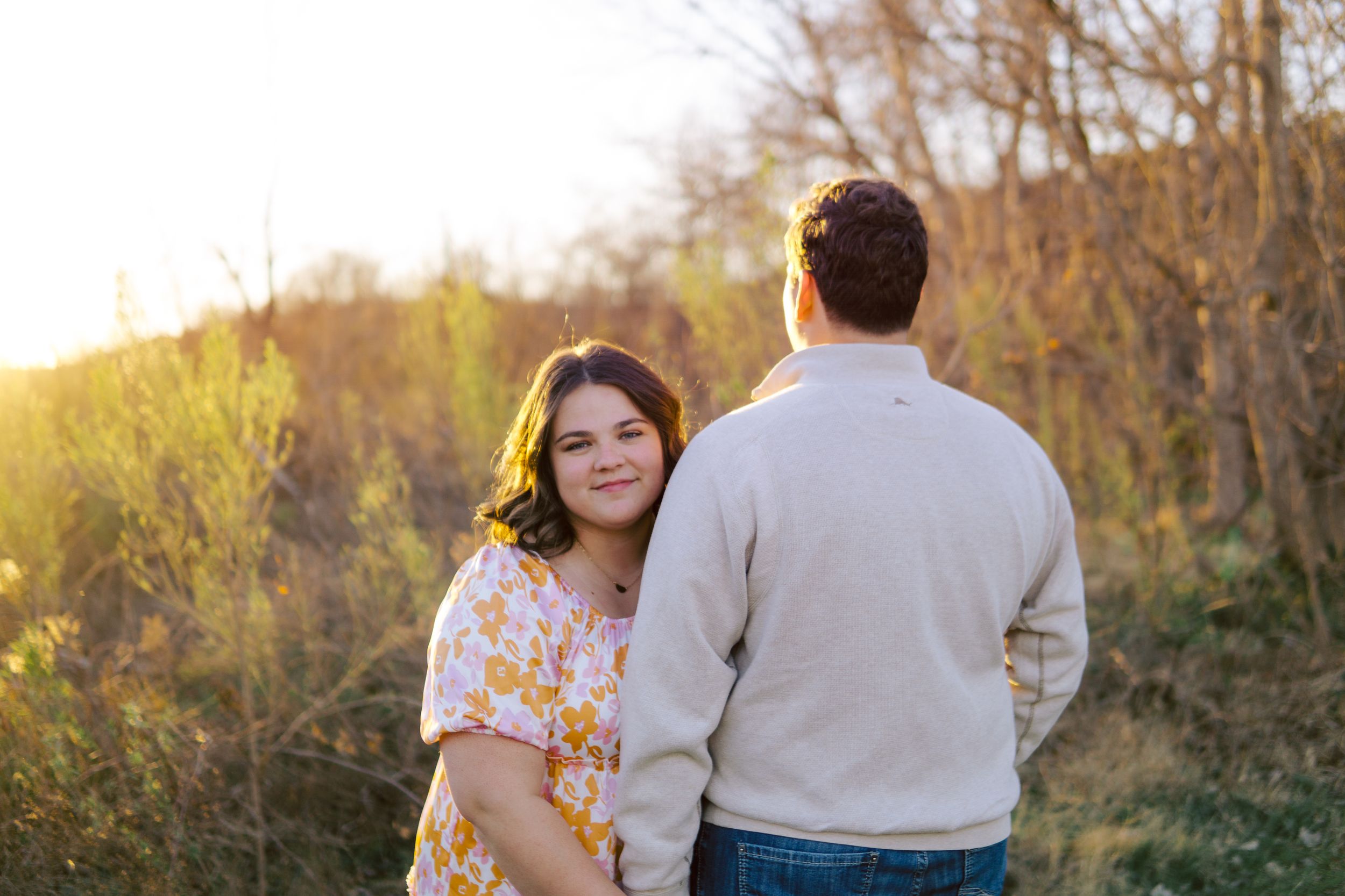Couple poses together outdoors during golden hour sunset with autumn trees in background.