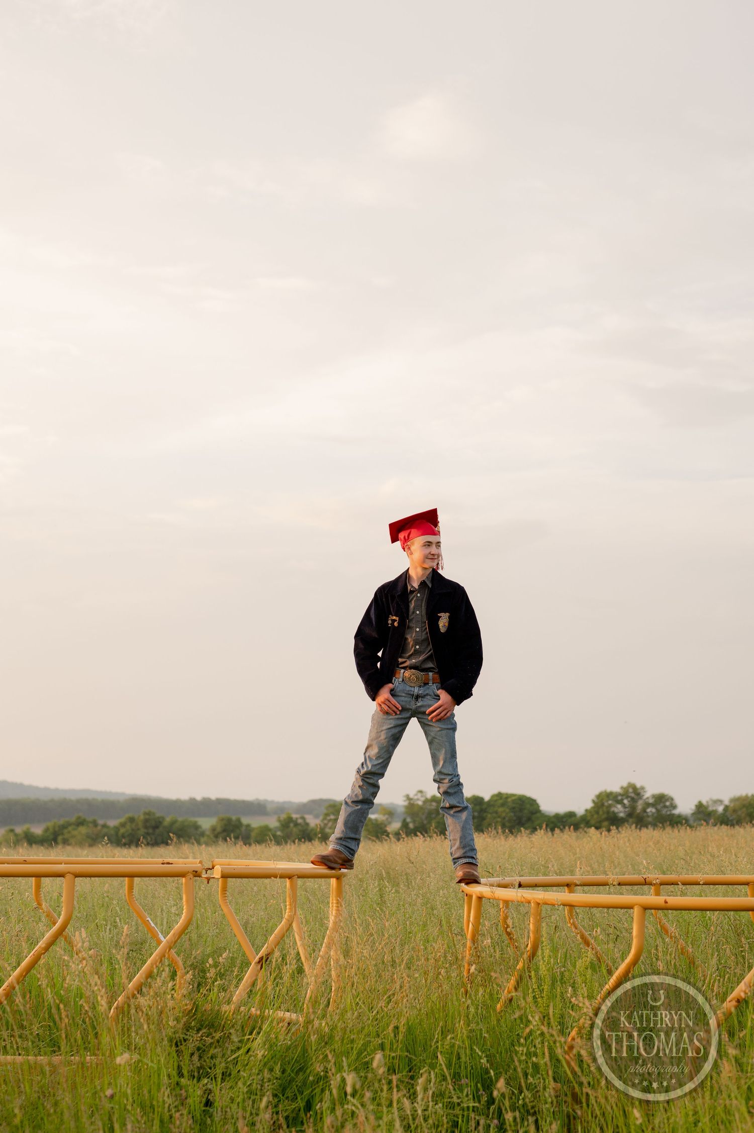 Student in FFA jacket and red cap poses confidently on wooden fence in rural field.