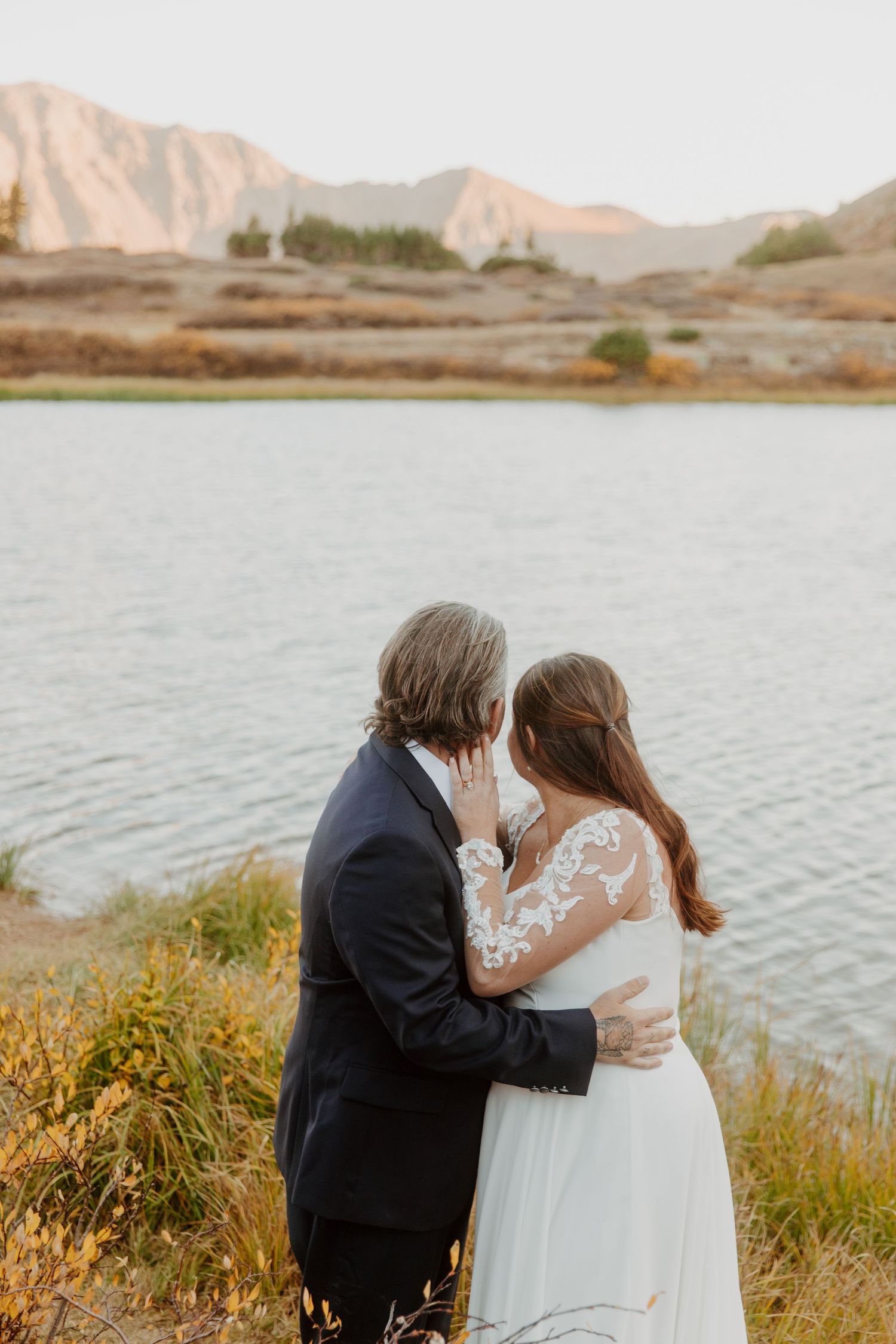 A couple in wedding attire embrace near a lake with desert mountains in the background at sunset.