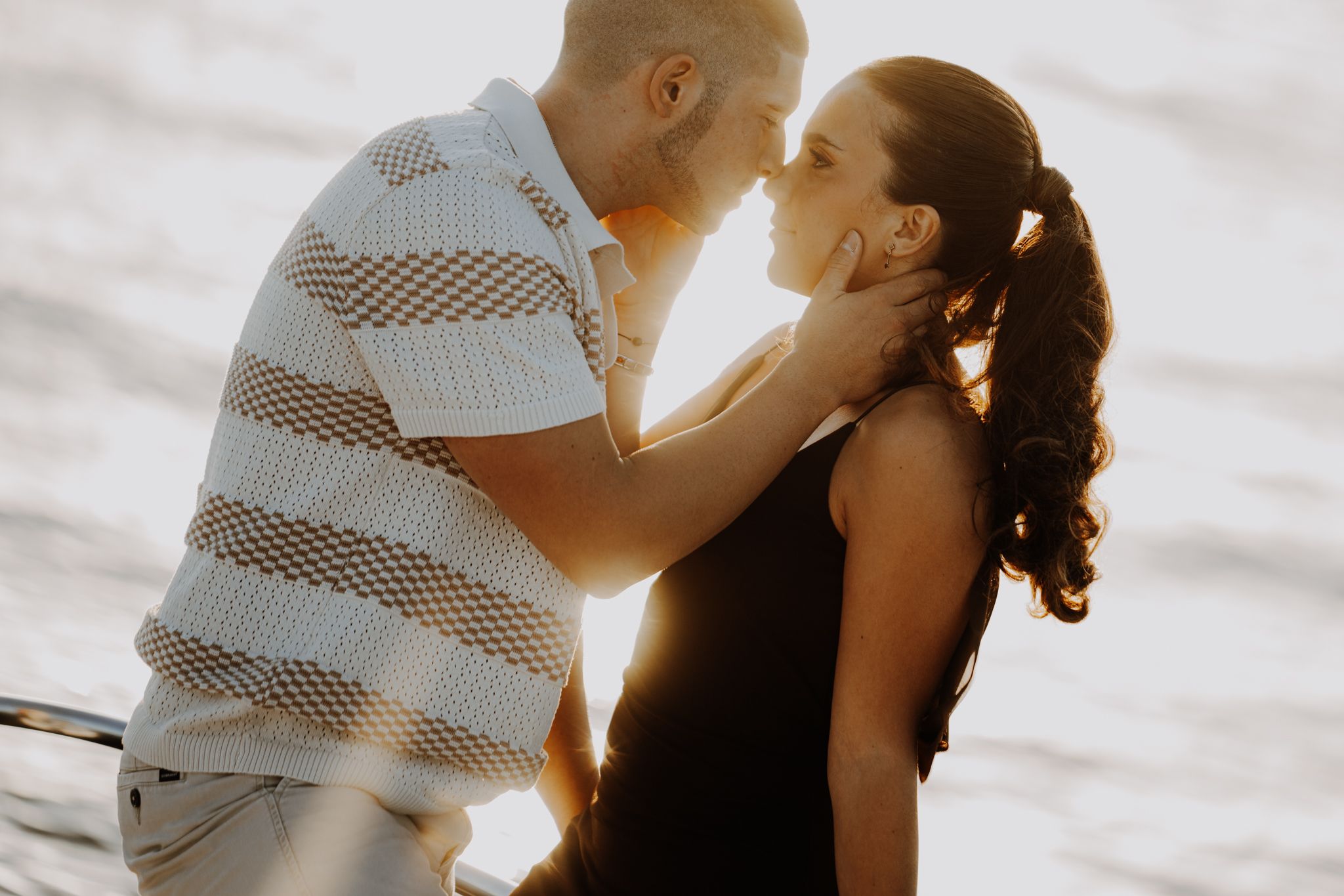 A couple shares a romantic kiss near the water at golden hour, the warm sunset light glowing behind them.