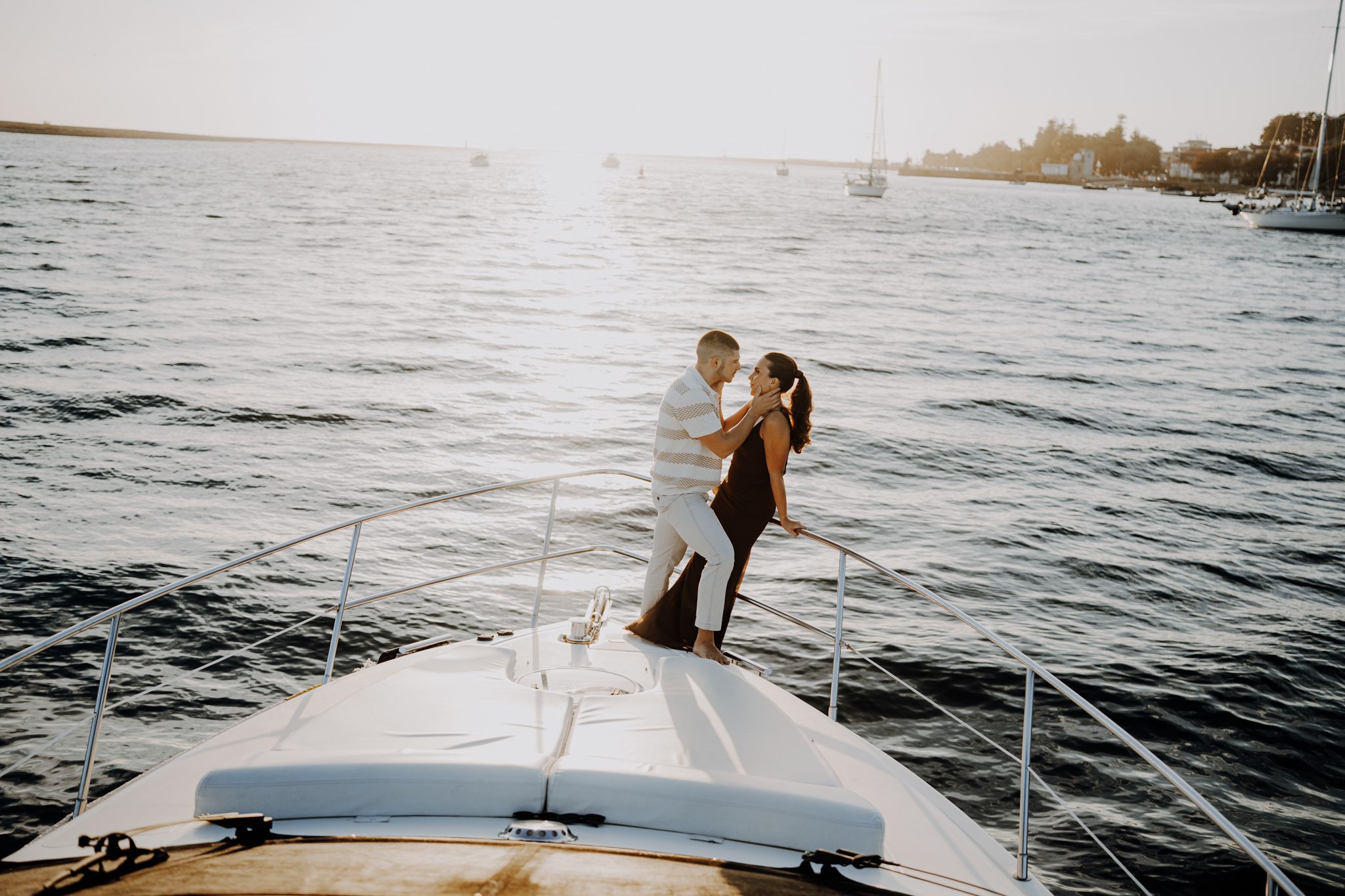 Couple embracing on the bow of a yacht at sunset with calm ocean waters and sailboats in the background.