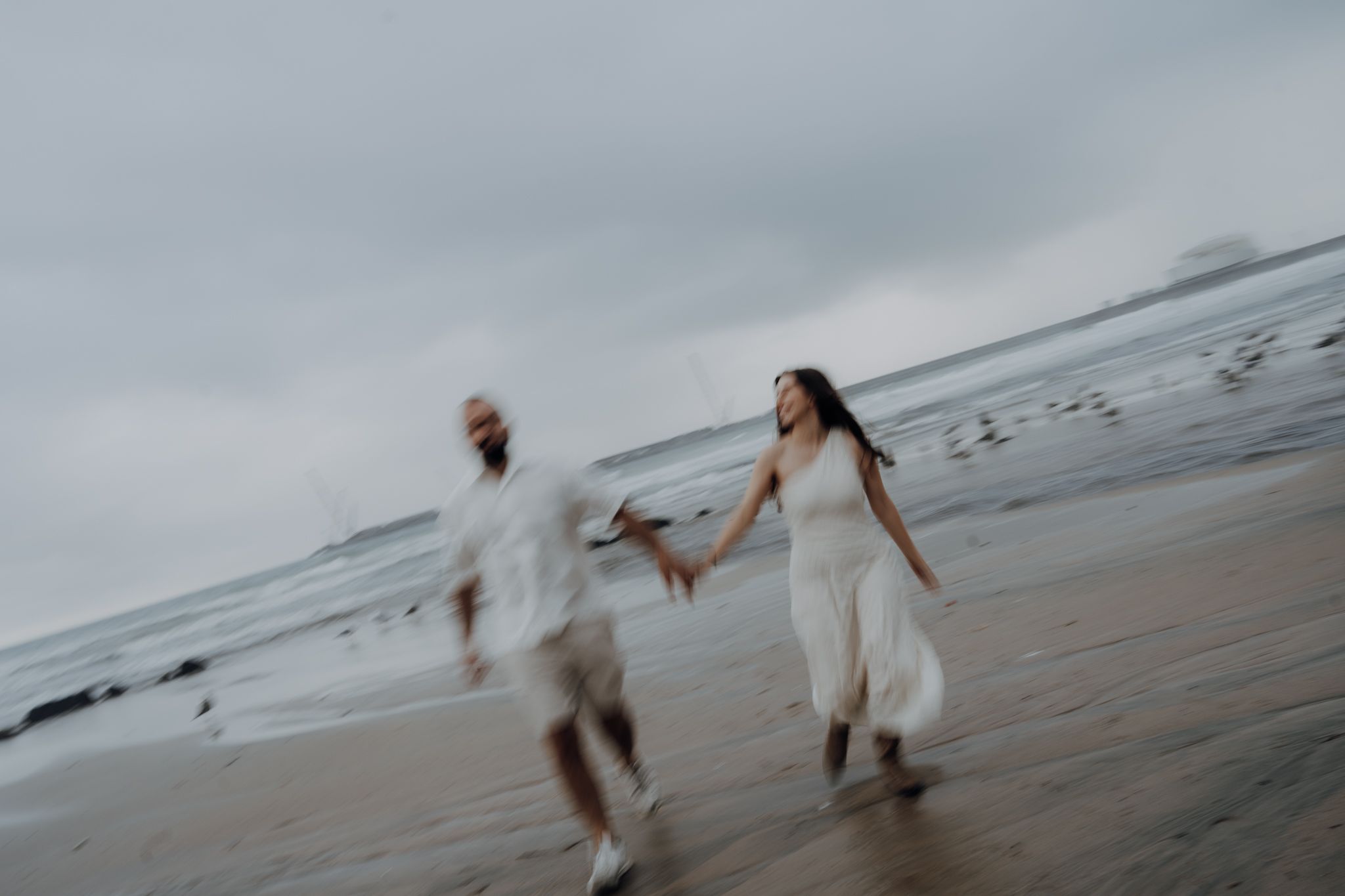 Blurred motion photo of a couple holding hands and running on a beach in white outfits on an overcast day.