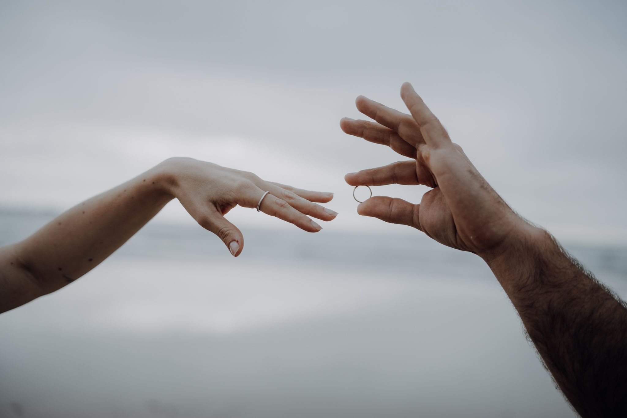 Two hands reaching toward each other against a cloudy sky, one holding a gold wedding ring.