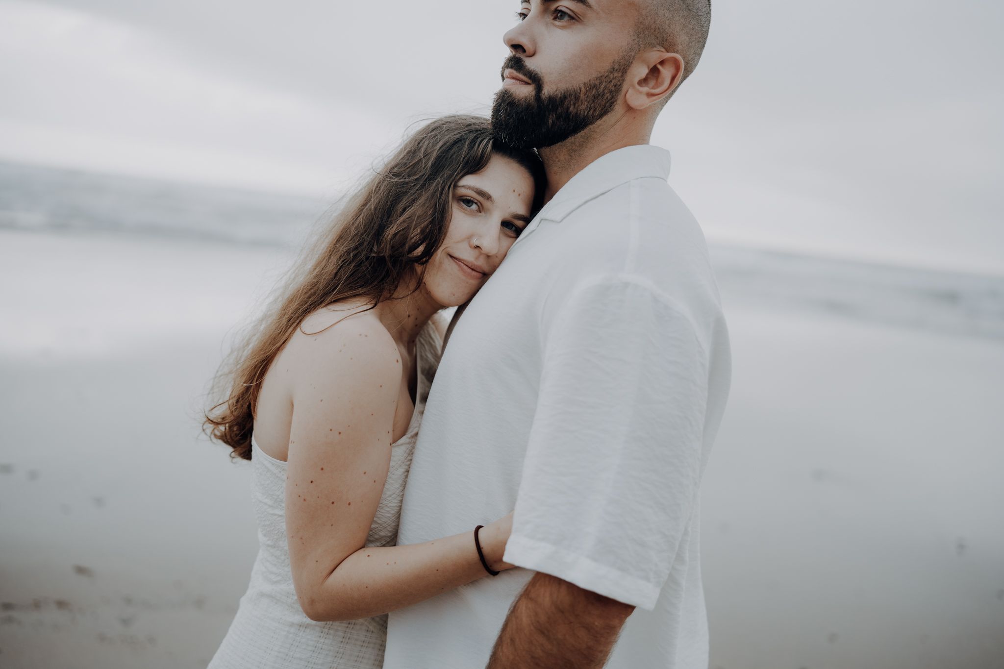 Couple embracing tenderly on a misty beach, woman in white dress leaning against man in white shirt.