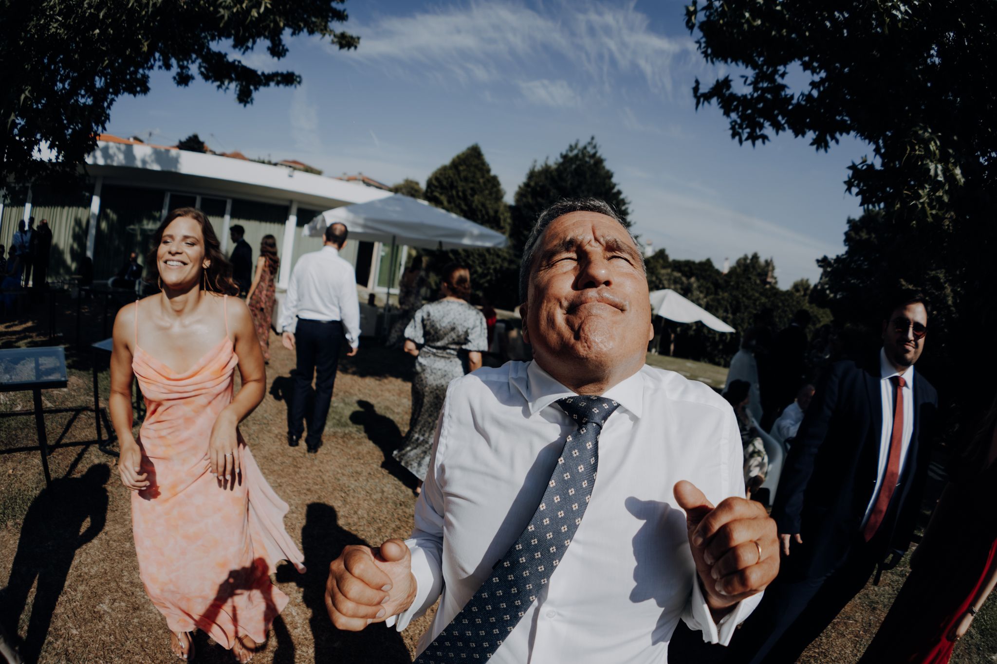 Man in suit looking up at outdoor wedding reception with guests socializing in background on sunny day.