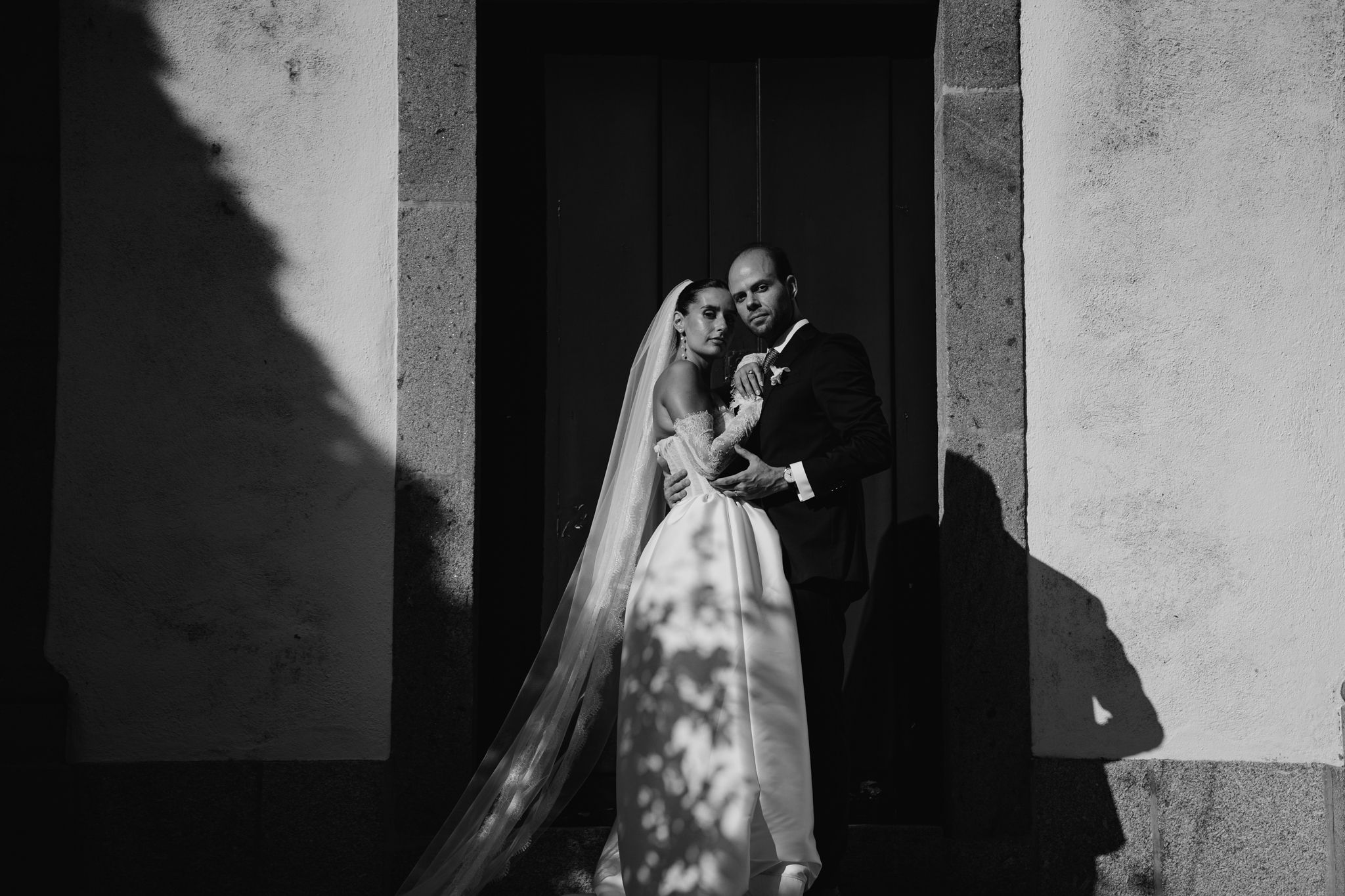 Black and white wedding photo of bride and groom embracing in dramatic light and shadow outside a stone building.